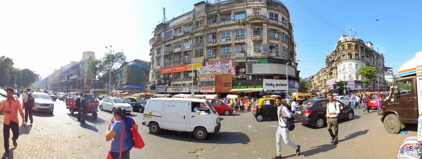 Wide panoramic street view of a busy Mumbai intersection showing colonial buildings, shops, vehicles including auto-rickshaws and cars, and pedestrians crossing under a clear blue sky