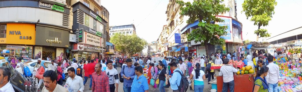 Crowded street market in Mumbai with pedestrians walking between street vendors selling colorful goods and multi-story buildings with shop signs including beauty palace and NIVA BAGS
