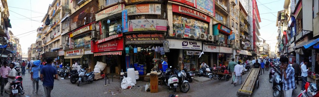 Wide street view of a busy Mumbai marketplace with multiple storefronts, people walking, and motorbikes parked along a commercial road lined with multi-story buildings