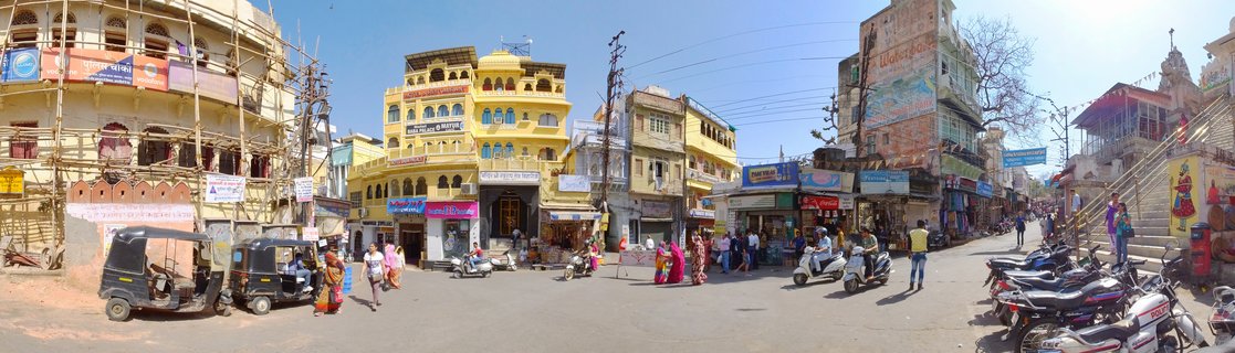 Wide panoramic street view of Udaipur, India showing colorful buildings, auto-rickshaws, scooters, pedestrians in saris, shops with Hindi signs, and temple steps under bright blue sky
