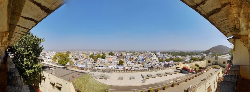 Wide panoramic photograph showing the city of Udaipur with white and blue buildings, stone architecture in foreground, clear blue sky above, and distant hills on the horizon