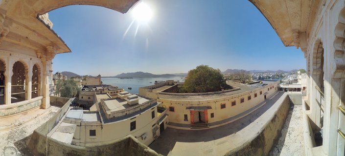 Panoramic view from City Palace courtyard showing cream-colored Rajasthani architecture with arched windows overlooking Lake Pichola with boats, distant mountains, and clear blue sky in Udaipur, India