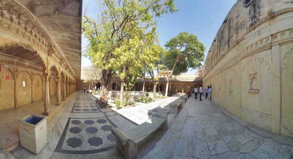 Panoramic view of a palace courtyard with carved stone columns, arched walkways, marble flooring, trees, and visitors at City Palace Udaipur India