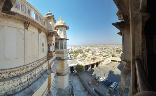Ornate cream-colored palace architecture with domed towers and carved stone balconies overlooking a courtyard with parked cars and the white cityscape of Udaipur, India under clear blue sky