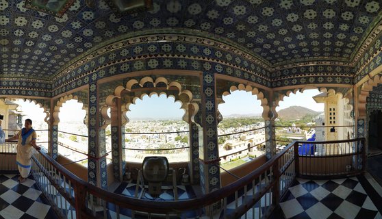 Ornate palace interior with blue patterned ceiling, arched openings, checkered floor, and woman in traditional dress overlooking Udaipur city