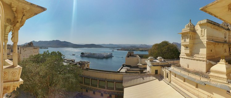 Panoramic view of the white Lake Palace on blue Lake Pichola water, framed by cream-colored ornate palace balconies and arches, with mountains visible in the distance under clear blue sky