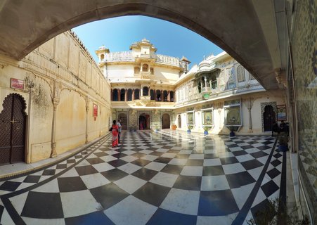 A palace courtyard with black and white checkered marble floor, cream-colored ornate buildings with balconies and peacock mosaics, framed by a stone archway, with visitors walking through the space under bright daylight