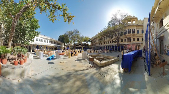 Wide panoramic view of a palace courtyard in Udaipur with cream-colored historic buildings, arched windows, event tables with white tablecloths, blue drapes, large trees, and clear blue sky