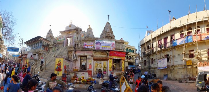 Wide street view showing a Hindu temple with multiple ornate spires, surrounded by shops with colorful signs, pedestrians walking, parked motorcycles, and a building covered in bamboo scaffolding under a clear blue sky