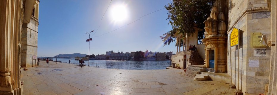 Wide panoramic view of Gangaur Ghat in Udaipur showing stone-paved lakeside promenade, traditional Rajasthani architecture, Lake Pichola with palace buildings, and distant hills under bright blue sky