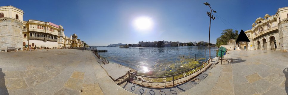 Wide panoramic photograph showing Lake Pichola in Udaipur with historic palace buildings on both sides, stone waterfront steps, calm blue water with distant island palace, bright sun in clear blue sky