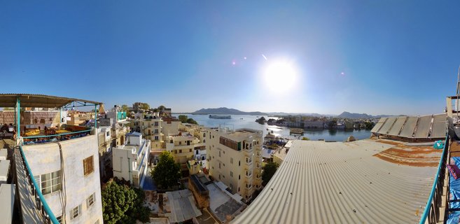 Aerial panoramic view of Udaipur showing Lake Pichola with Jag Mandir palace island, surrounded by cream-colored buildings and distant hills under a clear blue sky