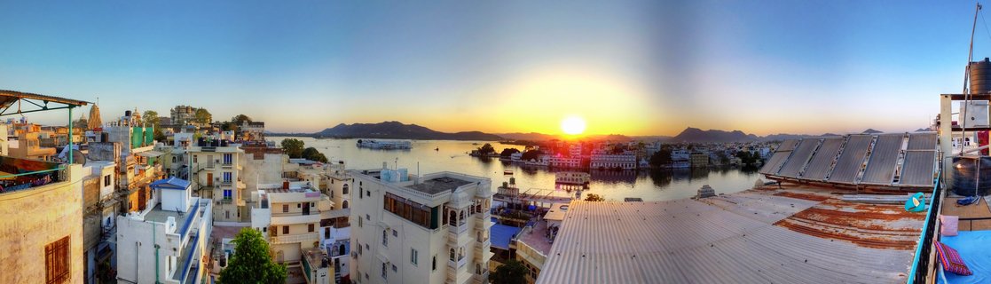 Panoramic sunset photograph of Lake Pichola in Udaipur, India, showing traditional white and cream buildings, solar panels on rooftops, calm lake water with boats, Jag Mandir palace island, and mountains silhouetted against golden-orange sky