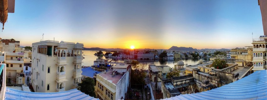 Panoramic sunset view over Lake Pichola in Udaipur showing Lake Palace Hotel, City Palace, traditional Rajasthani buildings, and Aravalli Mountains in the background