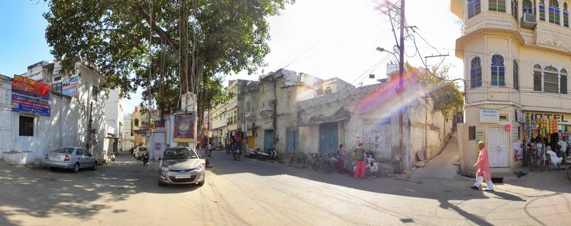 A panoramic street view in Udaipur showing traditional cream and white buildings, a large banyan tree, parked cars, pedestrians in colorful clothing, shops with merchandise displays, and overhead power lines under bright afternoon sunlight