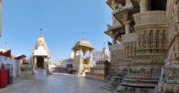 White stone Hindu temple courtyard with carved elephant friezes, ornate pillars, and shikhara spires against blue sky in Udaipur, India