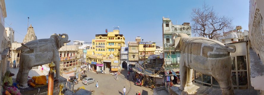 Elevated panoramic view showing two large stone elephant statues framing a busy street scene in Udaipur, India, with yellow buildings, pedestrians, vehicles, and a temple spire under clear blue sky