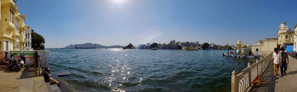 Panoramic view of Lake Pichola in Udaipur showing palace buildings on islands and waterfront, with people walking on stone ghats, motorcycles parked nearby, and mountains visible in the distance under bright blue sky