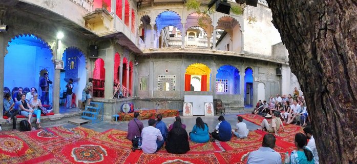 People seated on red patterned carpets in a courtyard with colorful arched doorways in blue, red, yellow and pink, watching a traditional Indian cultural performance in Udaipur, India