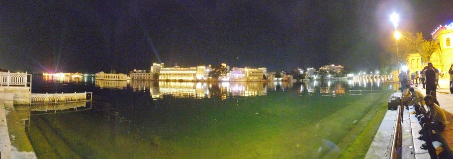 Nighttime panoramic view of illuminated Lake Palace and City Palace complex along Lake Pichola waterfront in Udaipur, Rajasthan, India with golden lights reflecting on dark calm water