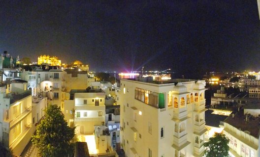 Night aerial view of Udaipur showing illuminated white buildings with traditional balconies, the glowing City Palace in the distance, and Lake Pichola reflecting city lights