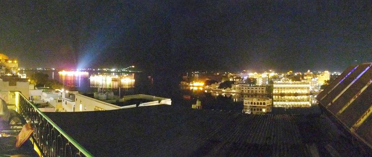 Nighttime panoramic view of illuminated palace buildings along Lake Pichola in Udaipur, India, with golden lights reflecting on dark water and traditional architecture visible from a rooftop vantage point