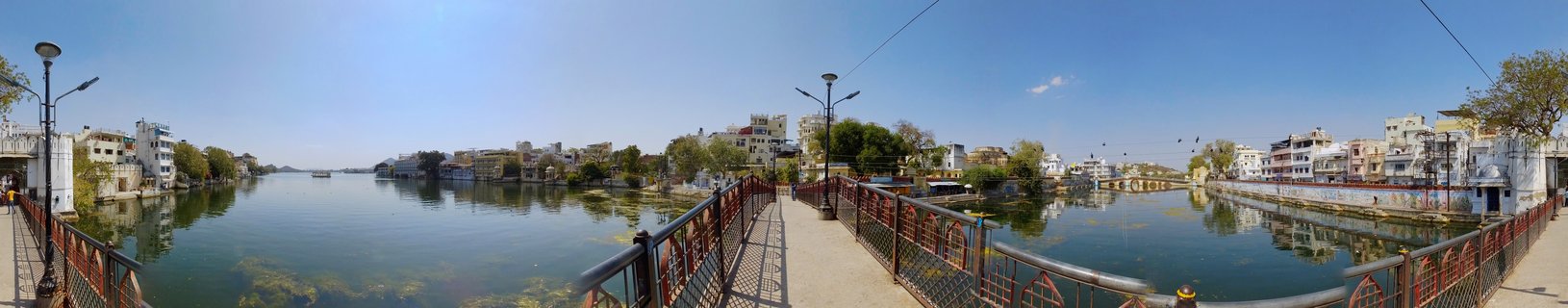 Panoramic view of Lake Pichola in Udaipur showing calm green-blue water with traditional buildings along the shoreline, a pedestrian bridge with metal railings in the foreground, and clear blue sky above.