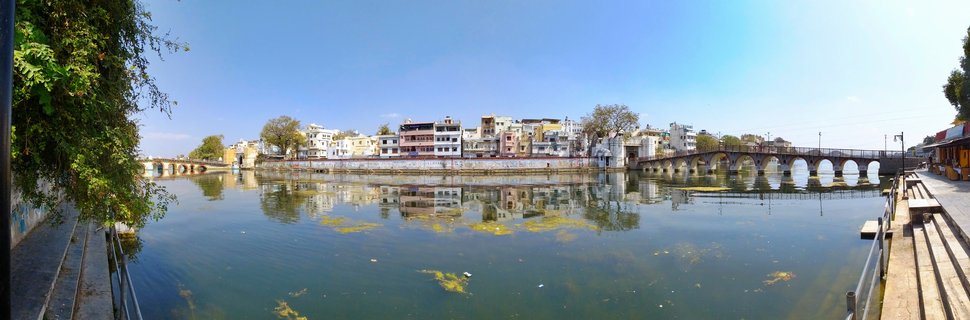 Panoramic view of colorful buildings and an arched stone bridge along a lake in Udaipur, India, with reflections visible in the water under a clear blue sky