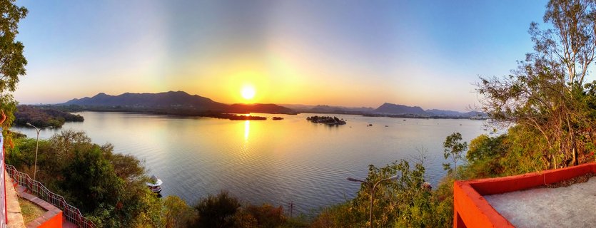 Sunset over a lake with small islands and mountains in the background, viewed from an orange terrace viewpoint with railings and trees framing the scene