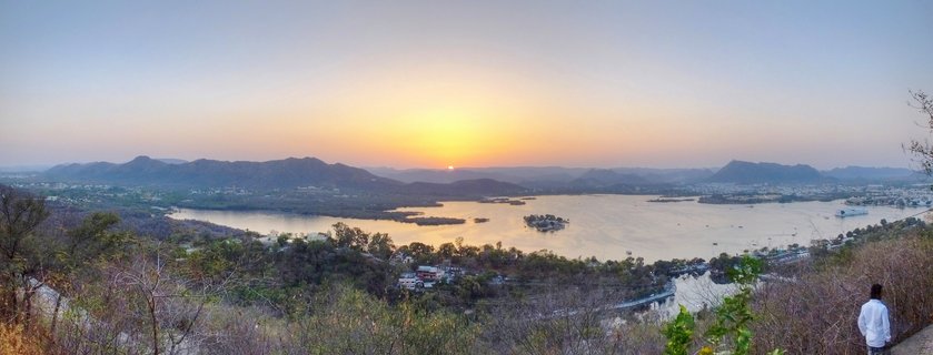 Panoramic landscape showing sunset over a large lake with islands, surrounded by mountains and vegetation in Udaipur, India