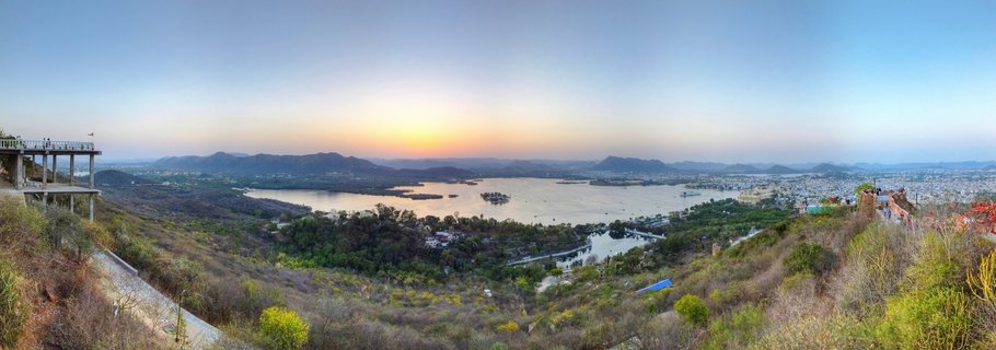 Panoramic landscape showing Lake Pichola in Udaipur at sunset with Jag Mandir island palace, surrounding hills, and city buildings under a gradient sky from blue to orange