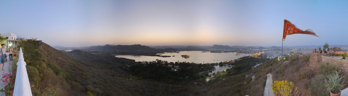 Wide panoramic photograph showing a large lake surrounded by hills and city buildings at sunset, viewed from an elevated hilltop position with architectural elements and a red flag visible in the foreground