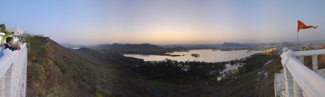 Panoramic sunset view showing Lake Pichola with palace islands, surrounded by hills and Udaipur city, viewed from an elevated white balustrade viewpoint with visitors and a saffron flag visible