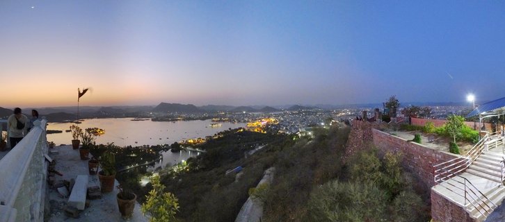 Panoramic evening view of Udaipur showing Lake Pichola with illuminated City Palace, city lights, and Aravalli hills under blue hour sky with crescent moon visible