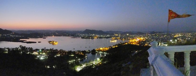 Panoramic twilight view of Udaipur showing illuminated Lake Palace on Lake Pichola with city lights, Aravalli hills in background, and white stone balustrade with red flag in foreground right
