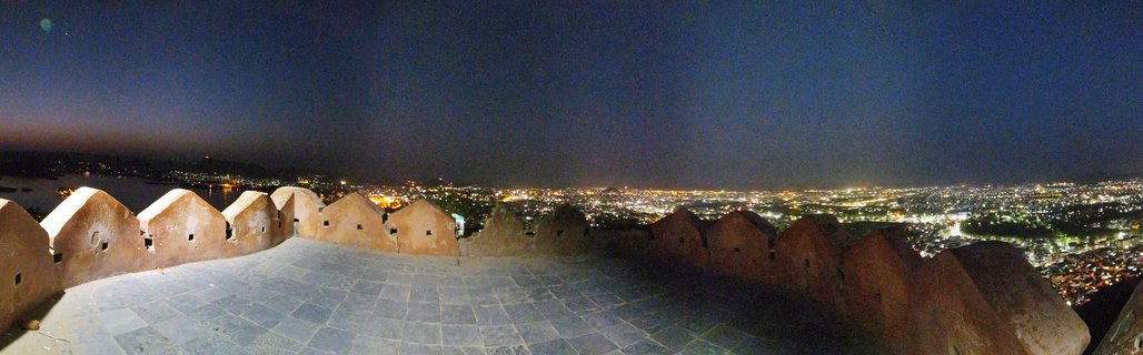 Night panoramic photograph showing traditional Indian palace fortress battlements in foreground with illuminated Udaipur city skyline and Lake Pichola visible in the distance under dark blue sky