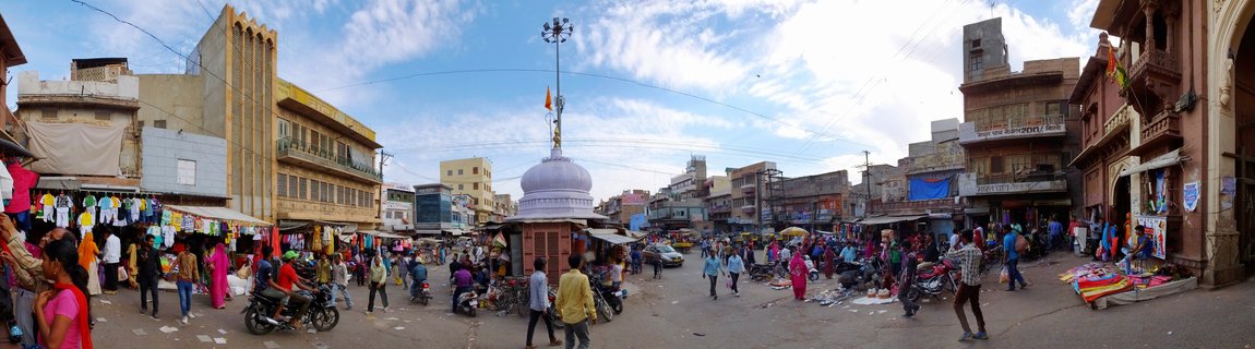 Wide panoramic view of a crowded street market in Jodhpur, India, with a white domed temple shrine in the center, people walking, motorcycles, colorful textiles for sale, and buildings on both sides under a blue sky with clouds.