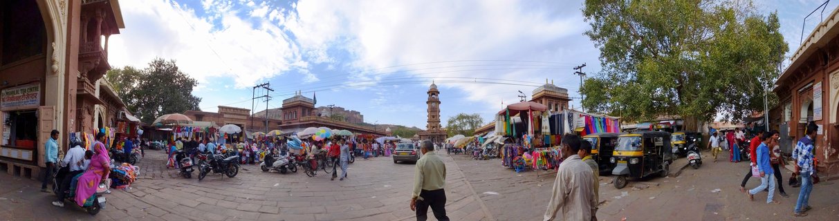 Wide panoramic view of a busy Indian marketplace with a clock tower in the center, surrounded by sandstone buildings, market stalls with colorful textiles, auto-rickshaws, motorcycles, and pedestrians in traditional clothing