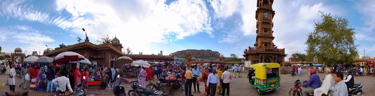 Busy outdoor market scene in Jodhpur India with a tall ornate clock tower, colorful market stalls with umbrellas, people in traditional clothing, auto-rickshaws and motorcycles under a blue sky with white clouds