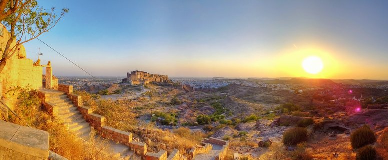 Panoramic sunset view of Mehrangarh Fort in Jodhpur, Rajasthan, India. The historic sandstone fortress sits atop a rocky hill in the center distance against a clear sky transitioning from blue to golden orange. The bright setting sun appears on the right. Stone steps and walls occupy the left foreground. Blue-painted buildings spread across the arid landscape below.