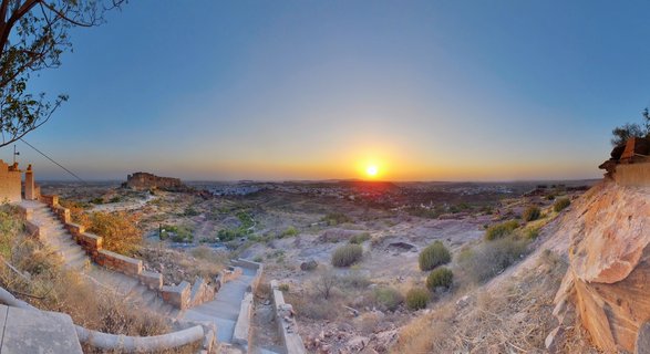 Panoramic landscape photograph of Mehrangarh Fort in Jodhpur, India at sunset. The historic sandstone fortress sits on a distant hill against a blue-to-orange gradient sky. Foreground shows rocky desert terrain with sparse vegetation, stone steps, and fortification walls. Golden hour lighting creates warm tones across the arid Rajasthan landscape.
