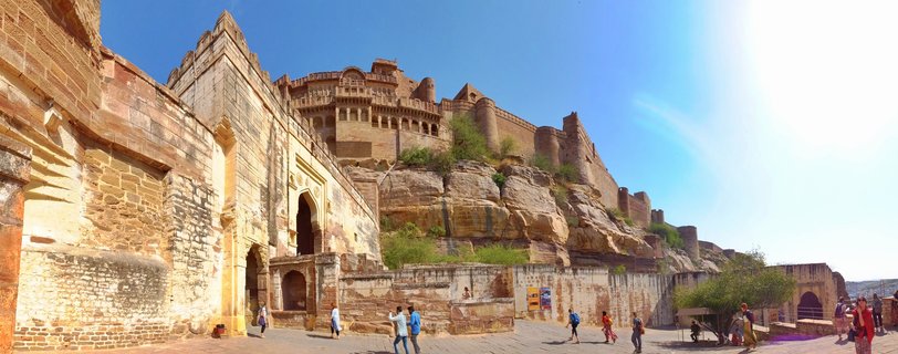 Golden sandstone fort built on rocky hill with tourists walking on paved courtyard below, clear blue sky above, Jaisalmer, India