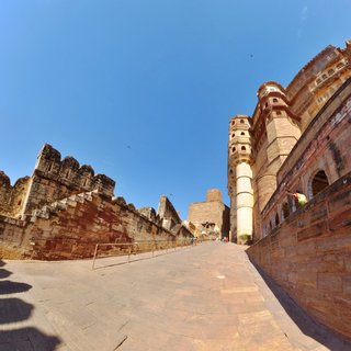 Stone pathway leading through Jaisalmer Fort with fortified walls on the left and ornate palace buildings with balconies on the right under blue sky