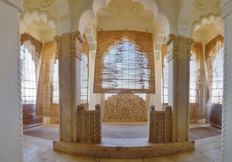 Ornate interior of Indian haveli with carved stone columns, arched openings, bamboo blinds, and decorative lattice screens