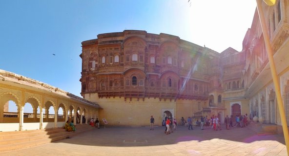 Wide courtyard of Jaisalmer Fort palace showing multi-story yellow sandstone building with ornate balconies, arched colonnade on left, and tourists walking on stone pavement under bright blue sky