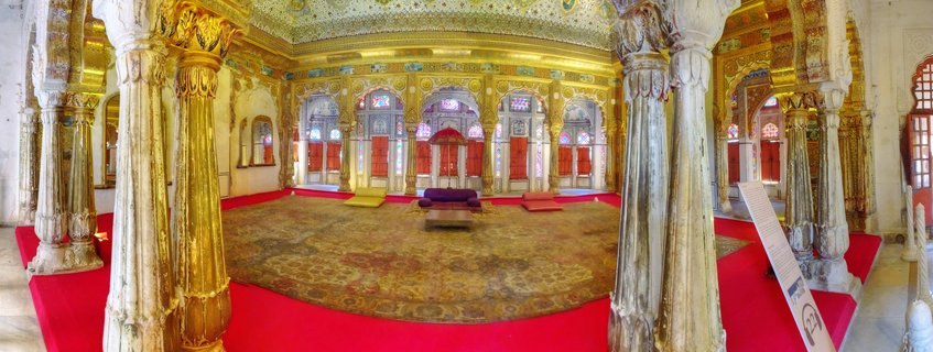 Ornate palace interior with golden carved columns, red carpet flooring, colorful stained glass windows, and traditional Indian architectural details in Jaisalmer, Rajasthan