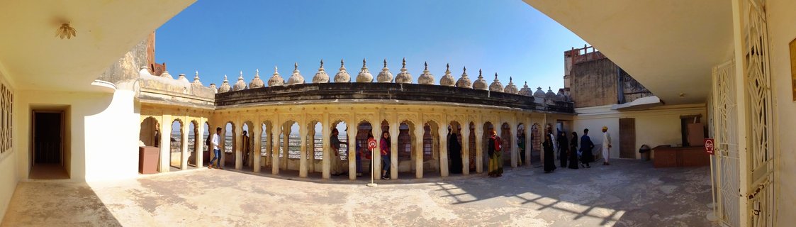Elevated view showing tourists in a stone courtyard at Jaisalmer Fort with ornate sandstone architecture featuring arched windows and the city of Jaisalmer visible in the background under clear sky