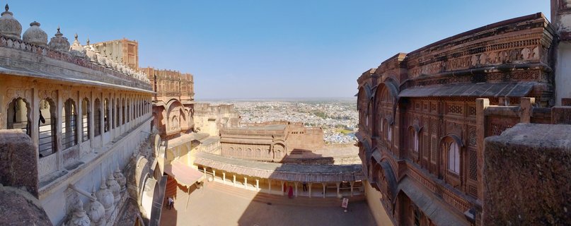 Panoramic view of Jaisalmer Fort courtyard showing golden sandstone buildings with ornate carved balconies, arched windows, and domed pavilions overlooking the city of Jaisalmer under clear blue sky