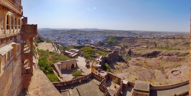 View from Mehrangarh Fort showing ornate sandstone architecture in foreground with blue-painted buildings of Jodhpur city and arid landscape extending to distant hills