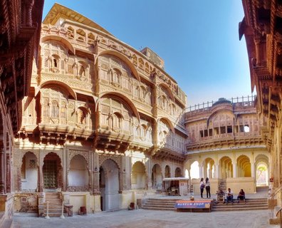 Wide view of an ornate palace courtyard with golden sandstone buildings featuring carved arches, balconies, and lattice windows surrounding a marble-paved open space with visitors walking through
